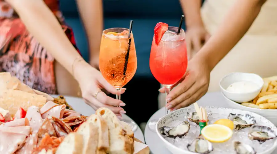 Hands reaching for colourful cocktail spritzes next to food platters