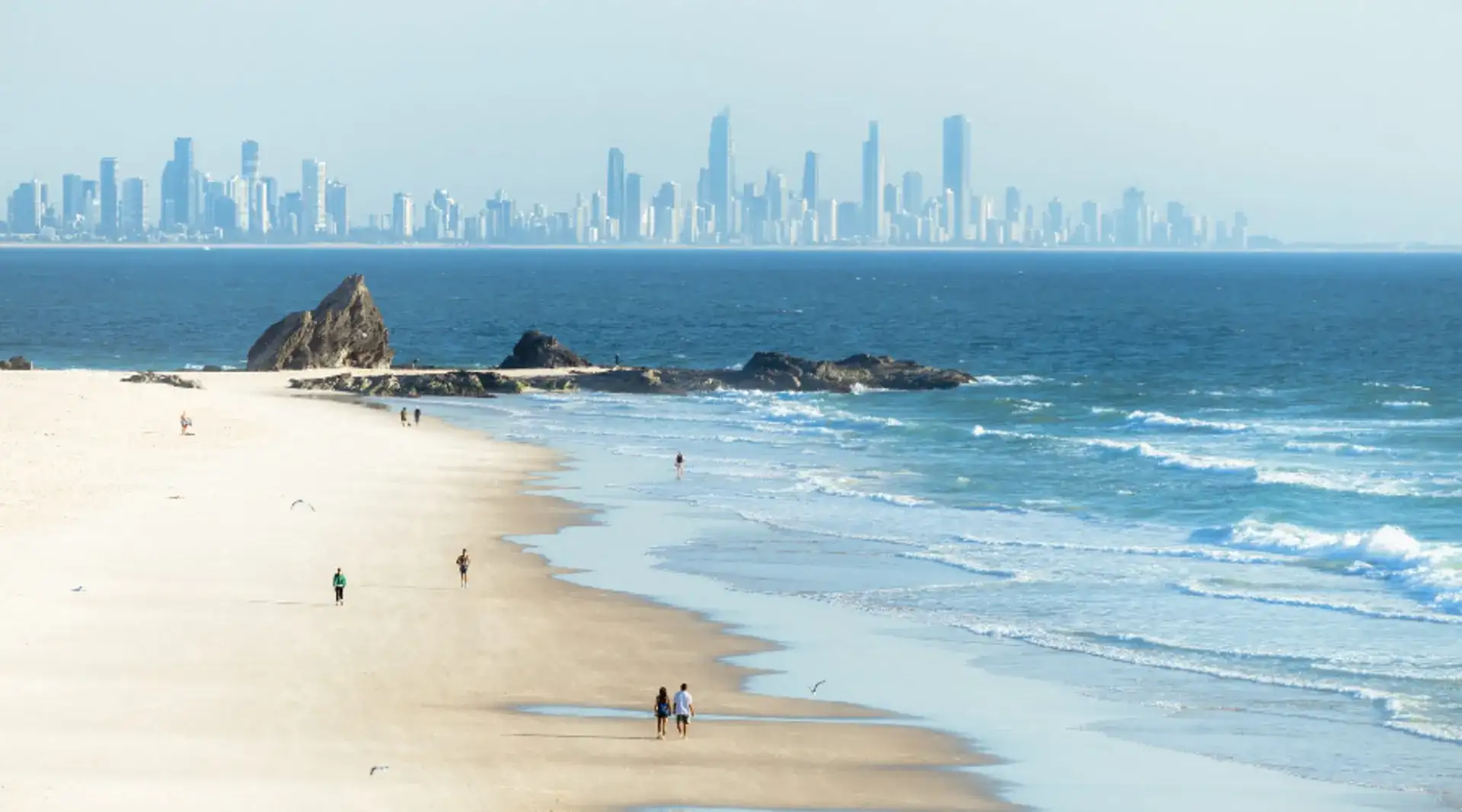 A couple walk along Currumbin Beach with city in backdrop