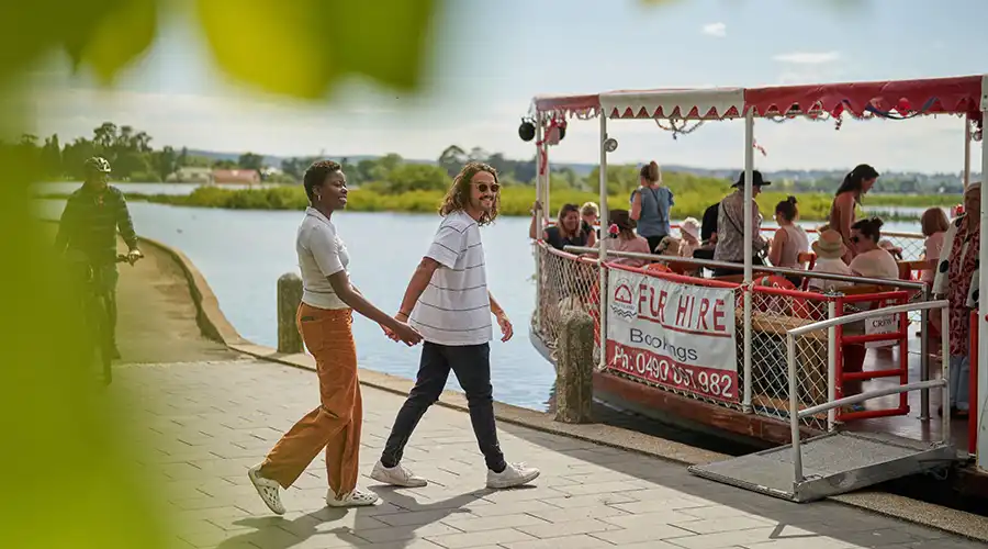 A couple holding hands walks along a lakeside path, passing by a boat filled with people enjoying a sunny day at the lake