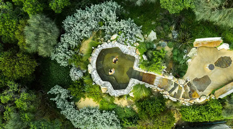 Aerial view of a natural hot spring nestled among lush greenery, with two people soaking in the serene, stone-lined pool