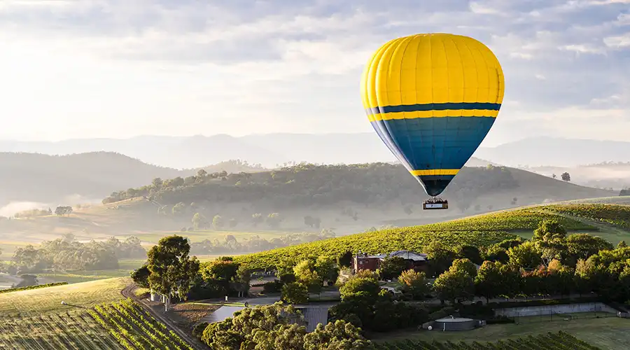 A vibrant hot air balloon floats peacefully over rolling green hills and vineyards in Yarra Valley, under a bright morning sky