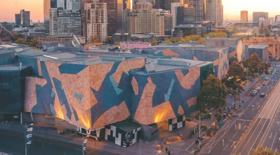 Evening shot of Federation Square in Melbourne, featuring geometric architectural designs, illuminated facades, and nearby bustling streets, representing a central hub of art and culture.