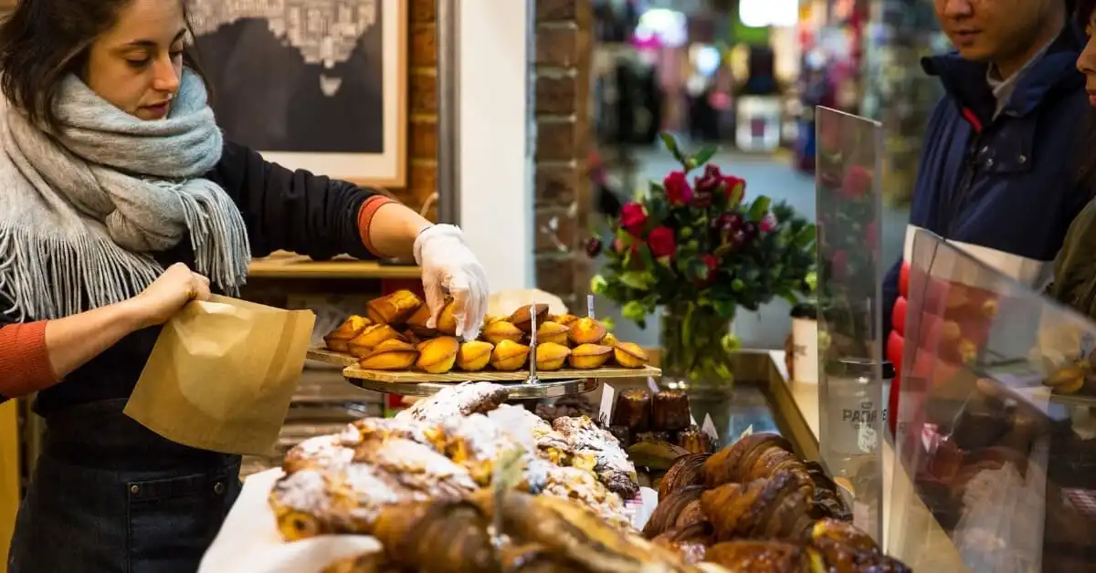 A woman in a scarf and apron carefully places freshly baked madeleines into a paper bag for customers at a pastry shop, with an array of pastries on display