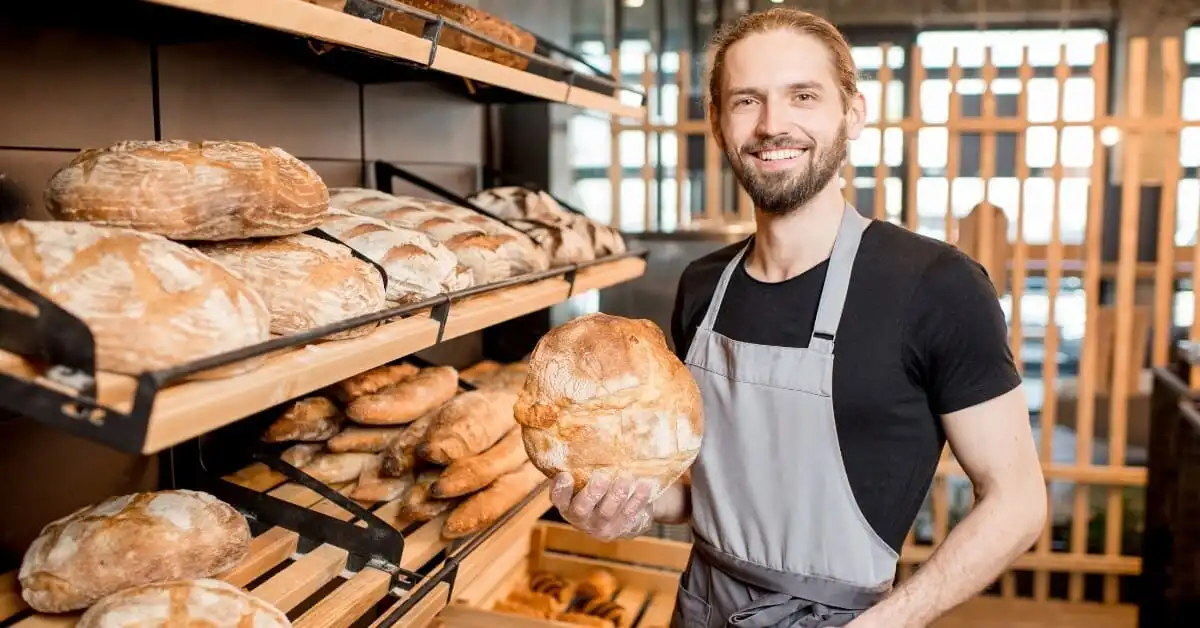 A smiling baker proudly holds a large, freshly baked artisan loaf of bread, surrounded by shelves filled with various types of bread in a bakery.