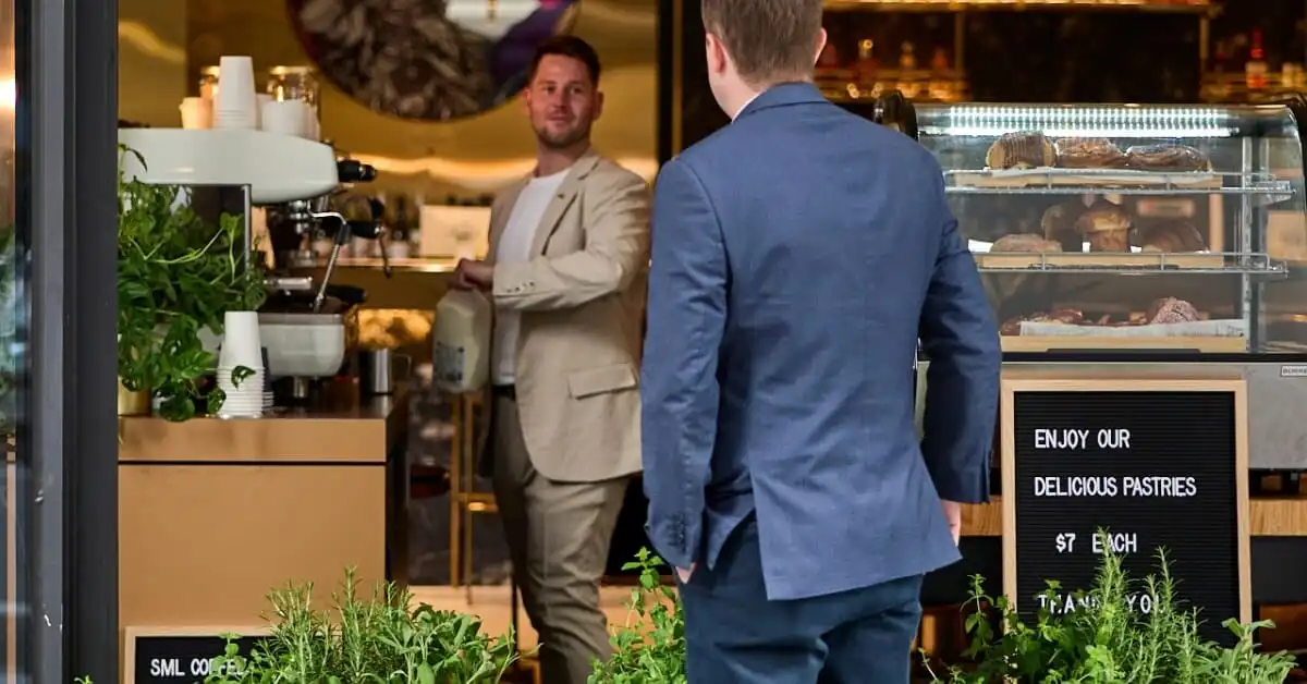 Two men in business attire engage in conversation outside a chic bakery café, with one man holding a carton of milk, and a display case filled with pastries inside