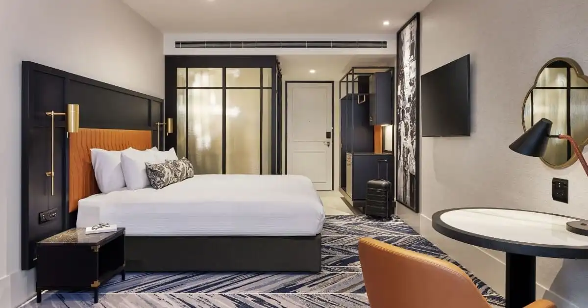 A stylish, modern hotel room with a large bed, sleek furnishings, and a well-lit, inviting atmosphere, featuring a patterned rug and a TV mounted on the wall