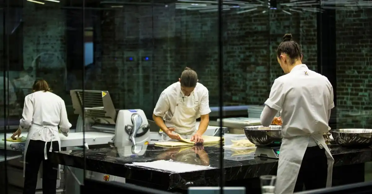 Three pastry chefs work diligently in a modern bakery kitchen, rolling and preparing dough on marble countertops, seen through a glass window