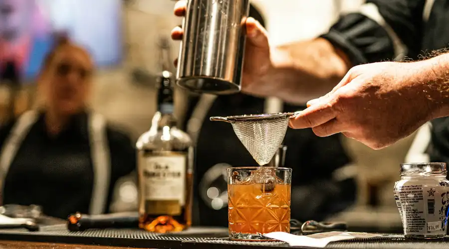 A bartender making a orange cocktail with whiskey bottle in the background, set against a warm background with people